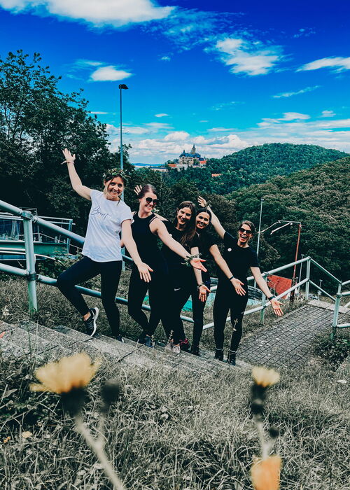 Fünf lachende Frauen posieren im Freien auf einer Treppe vor einer bewaldeten Hügellandschaft mit einem Schloss im Hintergrund unter blauem Himmel.